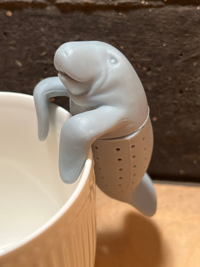 ManaTea infuser shaped like a manatee, hooks over a mug's rim. The perforated bottom holds loose-leaf tea, made from food-safe silicone for charming brewing.