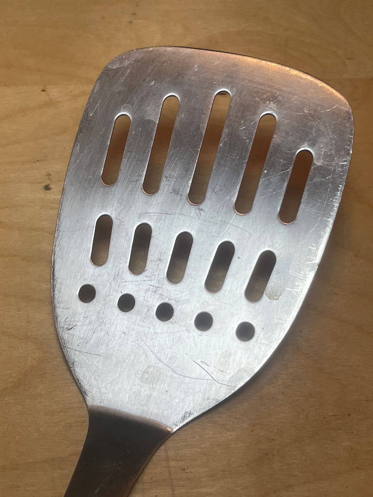 Wolfgang Puck stainless steel perforated spatula with bamboo handle resting on a wooden surface, showcasing its metal slots and ergonomic design.
