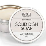 Small metal tin with Lovett Sundries label, containing solid dish soap puck. Zero waste, all-natural formula, lid slightly ajar, emphasizing eco-friendly cleaning.