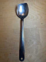 Blunt end serving spoon - Pan-Fit stainless steel, with a long handle and broad, shallow bowl, rests on a wooden surface, designed for versatile kitchen use.