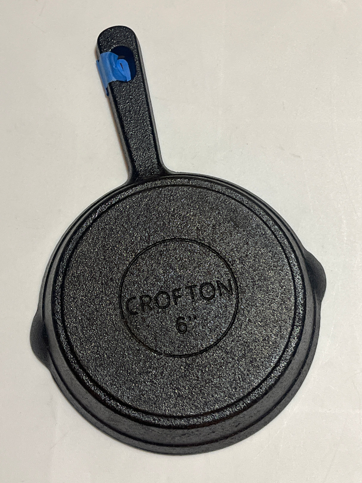 Crofton 6” teeny cast iron frying pan, underside view with Crofton 6″ label, handle wrapped in blue painter's tape, ideal for baking or frying.