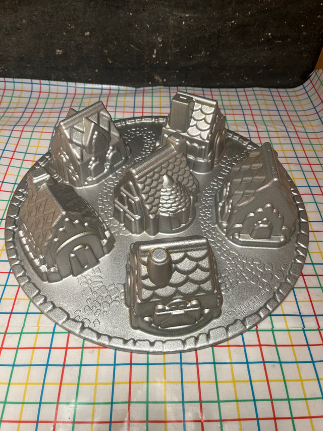 Cozy Village cast aluminum baking pan mold by Nordicware, featuring a ring of detailed house shapes on a grid-patterned tablecloth, ensuring even, nonstick baking.