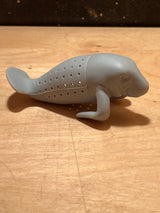 ManaTea infuser, a manatee-shaped silicone device, rests on a wooden surface, designed to hold loose tea leaves for steeping.