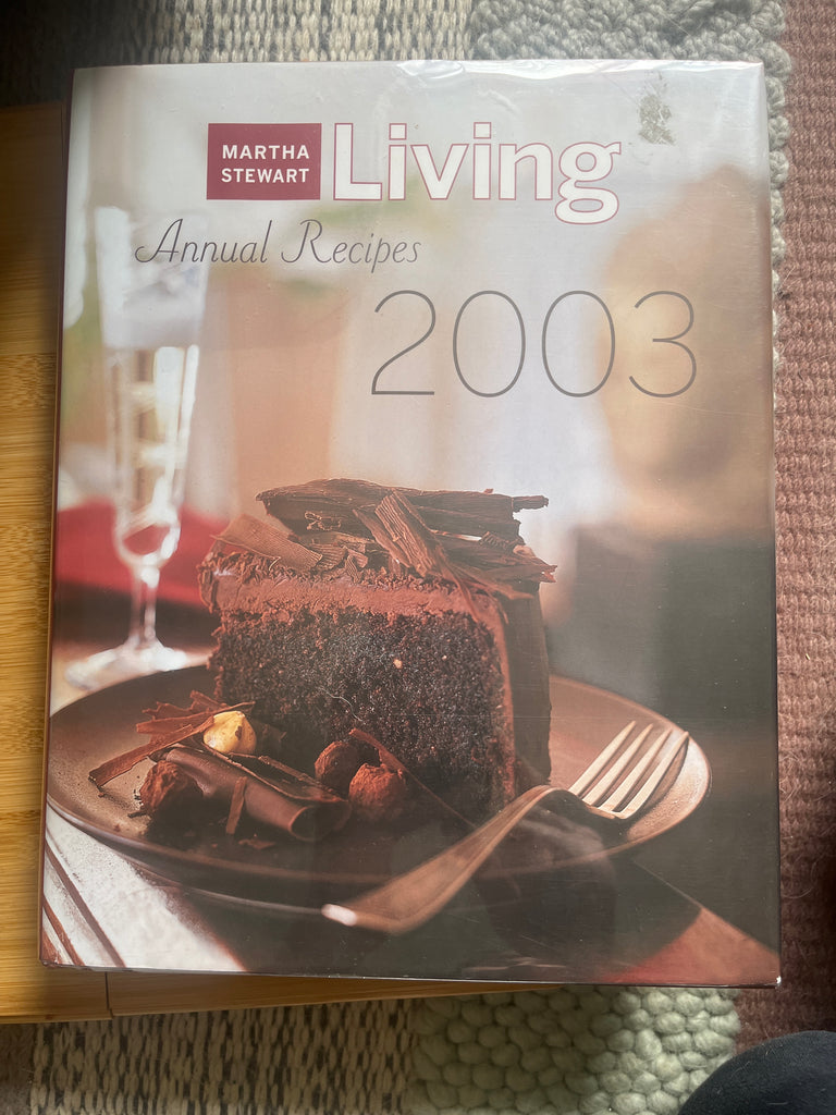 A book featuring Martha Stewart Living Annual Recipes 2003 with a slice of cake. Over 500 recipes and 96 photos organized by season, offering diverse culinary delights.