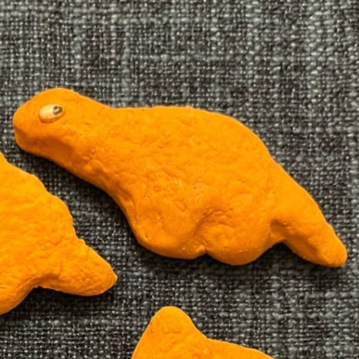 Orange diplodocous ceramic dino nuggie fridge magnet with small white and black eyes, placed on a textured gray fabric background