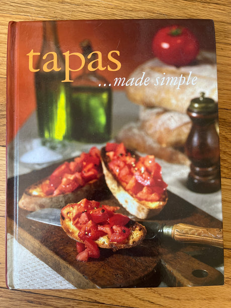 Tapas ... made simple cookbook on a table, featuring images of tomato bruschetta and other small dishes, highlighting delicious, easy-to-prepare Spanish recipes.