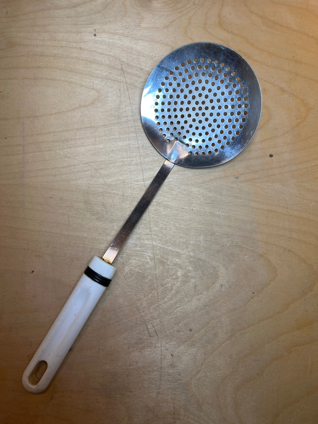 Round skimmer spoon - stainless steel with perforated head, features a long handle with white plastic grip, showing minor heat damage on the tip.