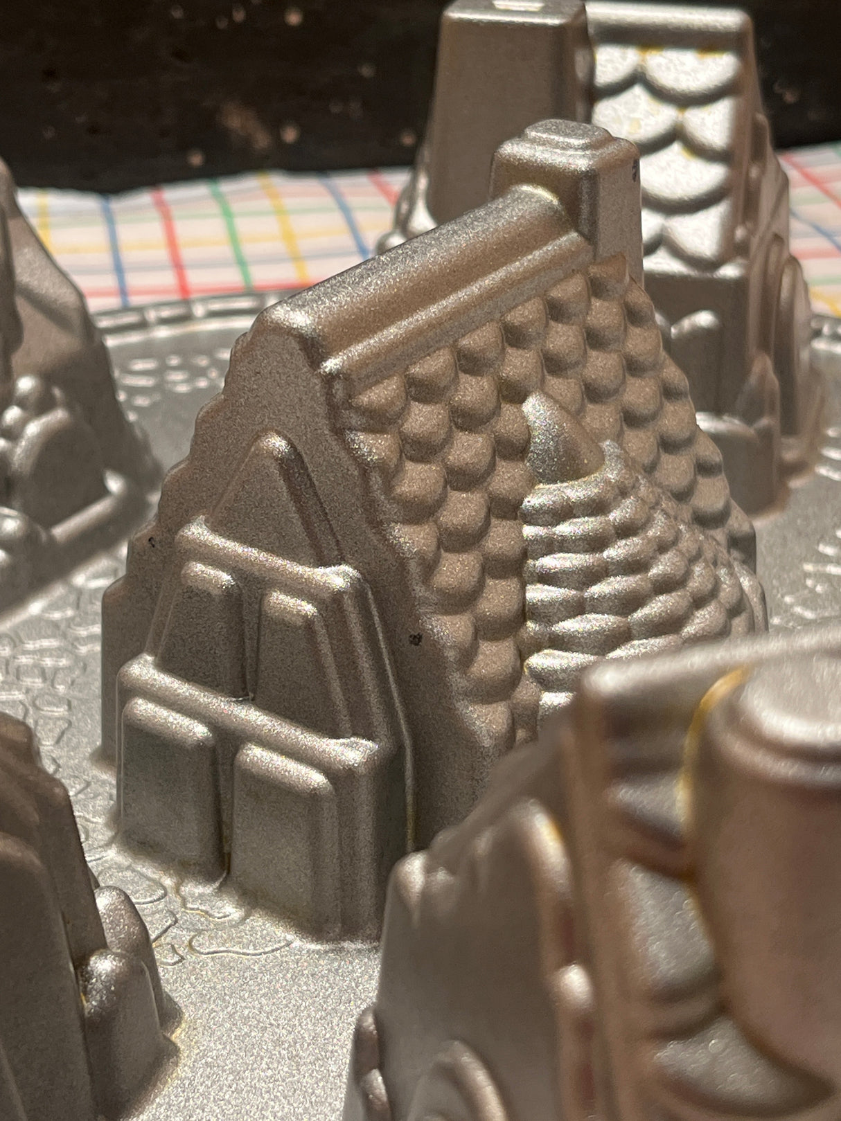 Cozy Village cast aluminum baking pan mold by Nordicware, featuring mountain and castle-tower shapes, ensuring uniform bakes with nonstick coating, visible against a plaid kitchen towel backdrop.