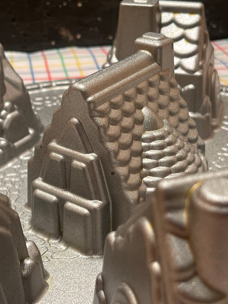 Cozy Village cast aluminum baking pan mold by Nordicware, featuring mountain and castle-tower shapes, ensuring uniform bakes with nonstick coating, visible against a plaid kitchen towel backdrop.