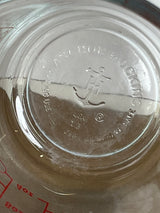 Underside view of an Anchor Hocking glass 2-cup measuring cup with handle, showing embossed anchor logo, safety markings, and partial red ounce-markings.