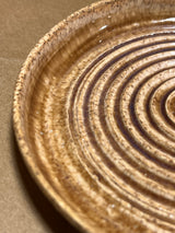 Handmade ceramic garlic grater spoon rest by Charles Lehman, featuring concentric ridges and a warm speckled glaze, displayed on a paper-covered surface.