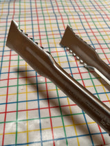 Large 16” VersaGrip Vollrath tongs with stainless steel scalloped ends and black Kool-Touch® handle, resting on a checked tablecloth.