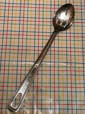 Extra long slotted basting spoon - JR stainless steel, featuring a durable, easy-to-clean long handle, resting on a tabletop with a grid-patterned tablecloth.