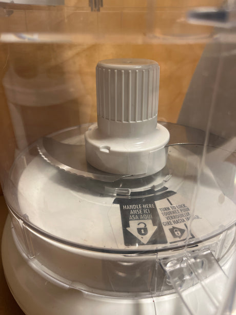 Hamilton Beach Chef Prep 70700 7-cup food processor close-up showing clear plastic lid with safety instructions, central drive shaft, and stainless-steel blade.