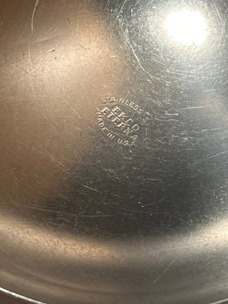 Stainless steel ladle with wood handle - EKCO Eterna, featuring a wide base for easy scooping, shown over a stainless steel pan bottom with logo and stamp.
