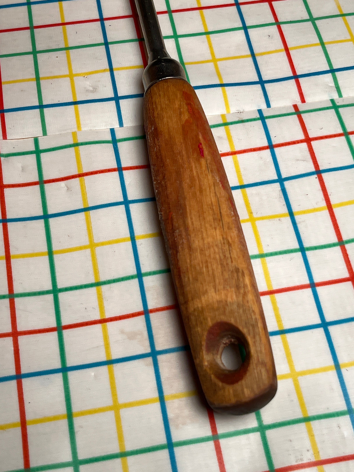 Vintage flat thin steel slotted spatula with wood handle, featuring a metal ferrule and hang hole, resting on a multicolored grid-patterned surface.