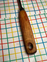 Vintage flat thin steel slotted spatula with wood handle, featuring a metal ferrule and hang hole, resting on a multicolored grid-patterned surface.