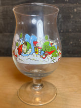 Huvel Belgian beer glass with limited edition 2024 JT Samuels illustration of hop cones, stylized characters, and foam, on a wooden surface.