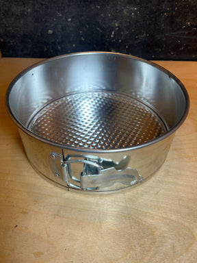Springform cake pan - light tin 8” diameter, featuring a round metal design with a latch on the side, ideal for easy cake release on a wooden surface.