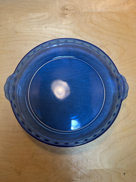 Pyrex 229 cobalt blue glass pie/casserole dish, round and translucent with fluted edges, small handles, and deep sides, shown upside-down on a wood tabletop.