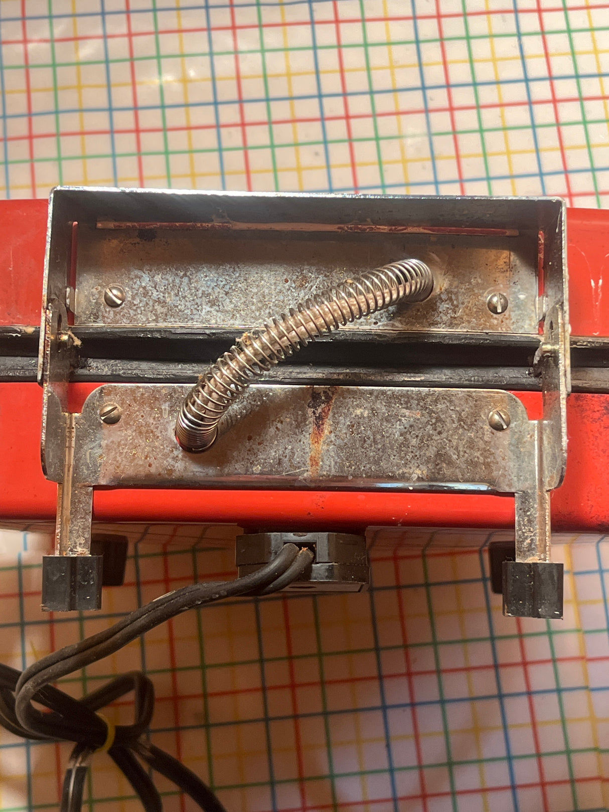 Underside view of Waffle iron - red vintage Wards Signature BE-46029, showing metal bracket, spring-wrapped conduit, black standoffs, and bundled electrical cables on a patterned surface.