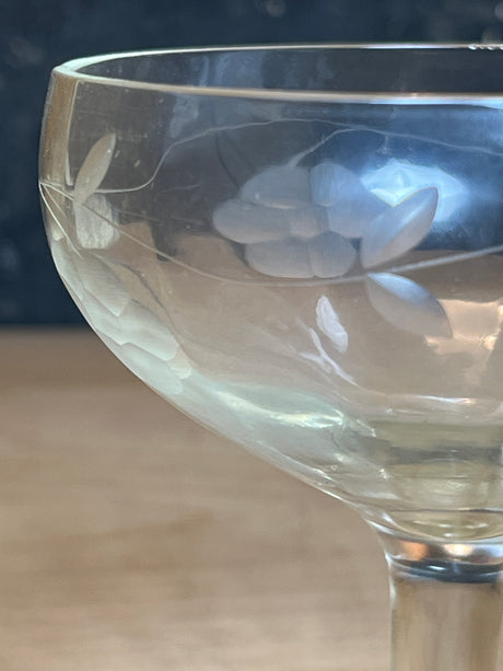 Close-up of a Princess House Heritage Pattern etched glass wine goblet, showcasing a grape-and-leaf motif on the bowl, resting on a wooden surface.