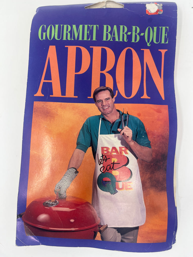 A man in a Let's Eat! Gourmet Bar-B-Que apron, holding tongs and a grill tool, ready for a BBQ. Original packaging, never worn.
