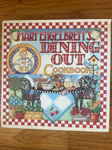 Mary Engelbreit's Dining Out Cookbook cover featuring a table with a cookbook, a cartoon woman holding a spatula, and various food items.