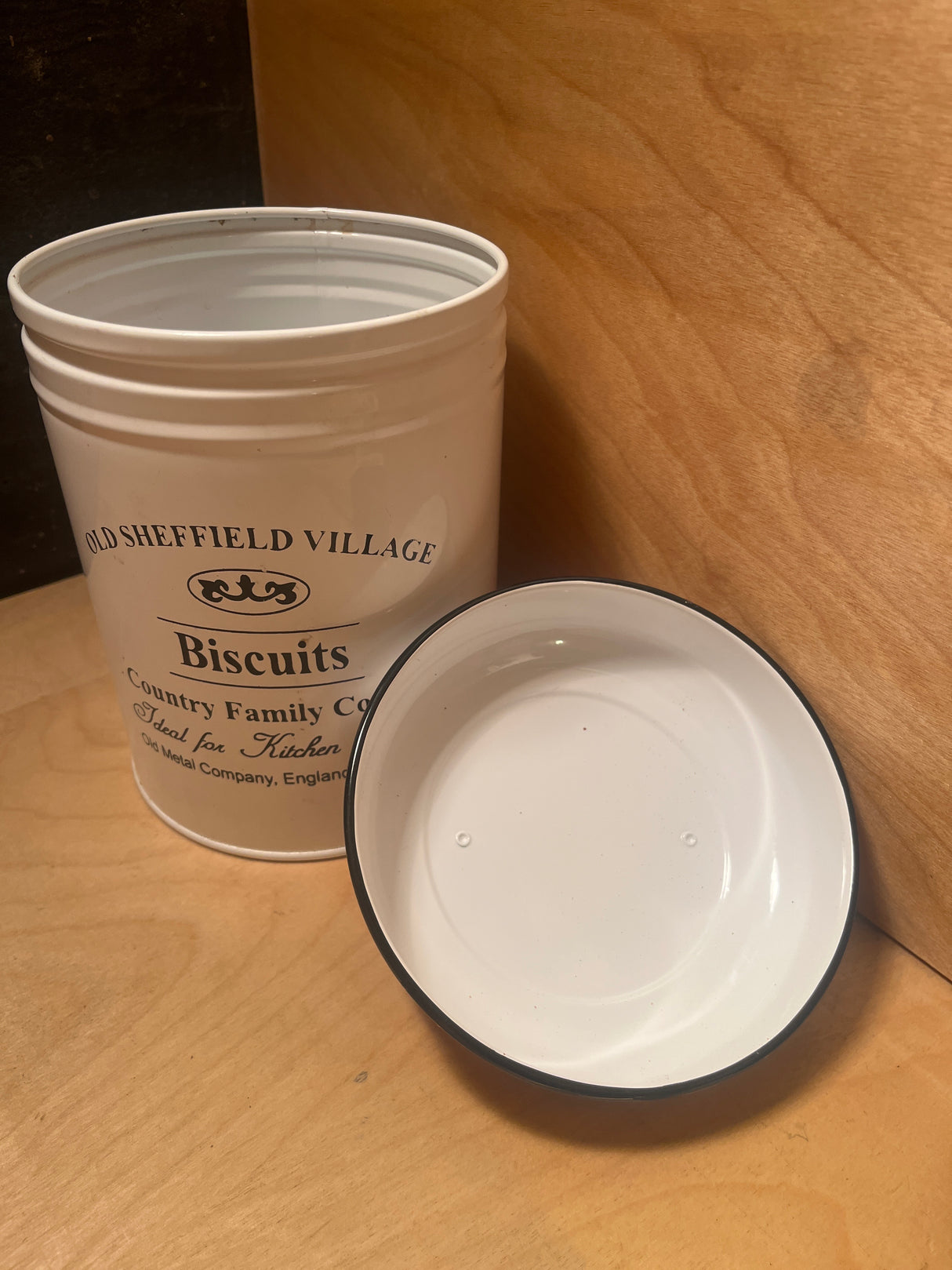 Old Sheffield Village Country Family Cottage Biscuits tin, white metal, cylindrical, open with lid beside it on wooden surface, ideal for vintage kitchen decor.