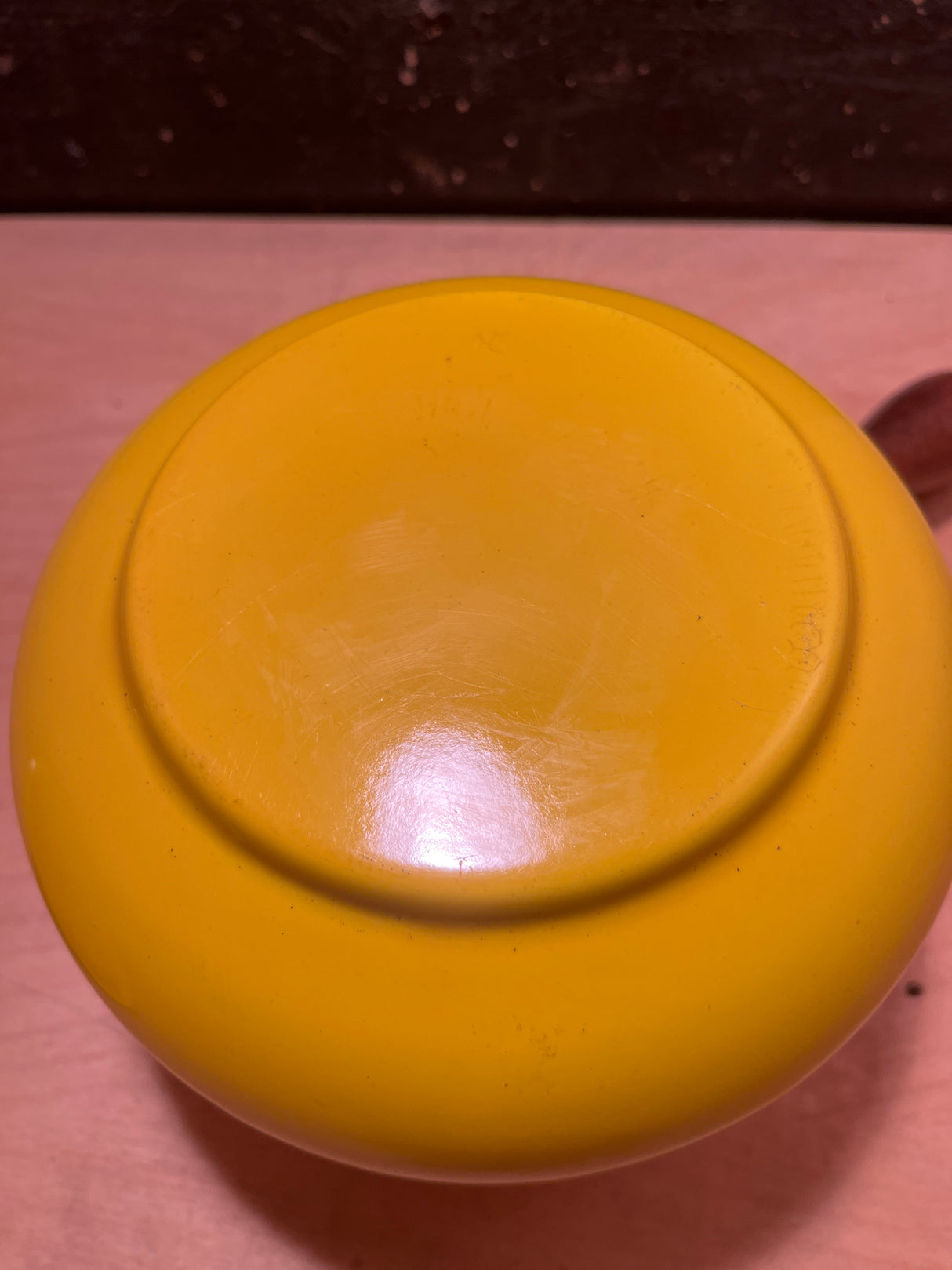 Vintage enamel sunshine yellow fondue pot with visible foot ring, flipped upside-down on wooden surface; glossy finish shows surface scratches, complemented by wood handles.
