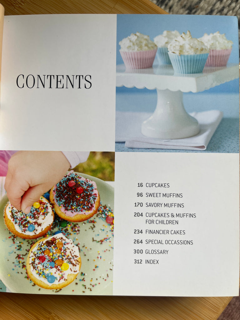 A cookbook featuring 100 cupcake and muffin recipes, displayed with images of cupcakes with various frostings and sprinkles on a white stand.