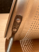 Close-up of a vintage aluminum Cone sieve strainer DENTED, showing perforated wall, riveted handle bracket with rust, and minor dents in cone tip.