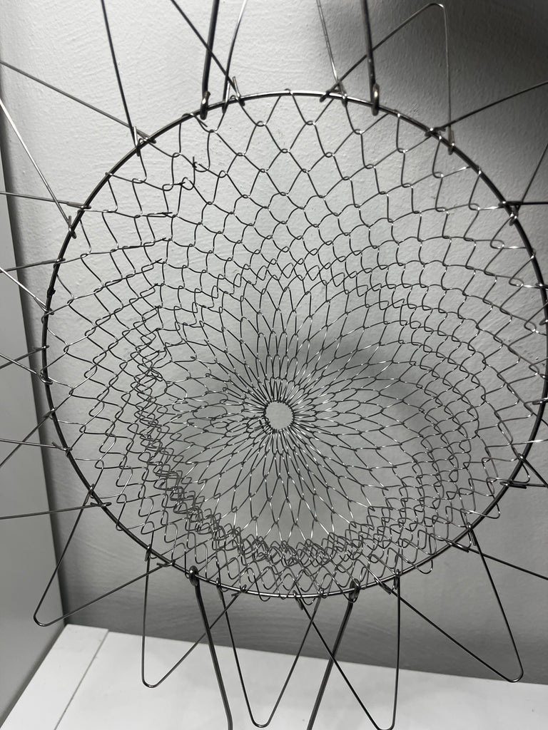 Circular wire mesh basket for frying and steaming food, with spiral design and metal rods unfolded.