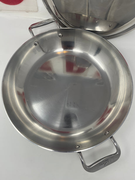 Close-up of an All-Clad stainless steel paella braise pan, featuring double handles and a domed lid, highlighting its sleek metal surface.