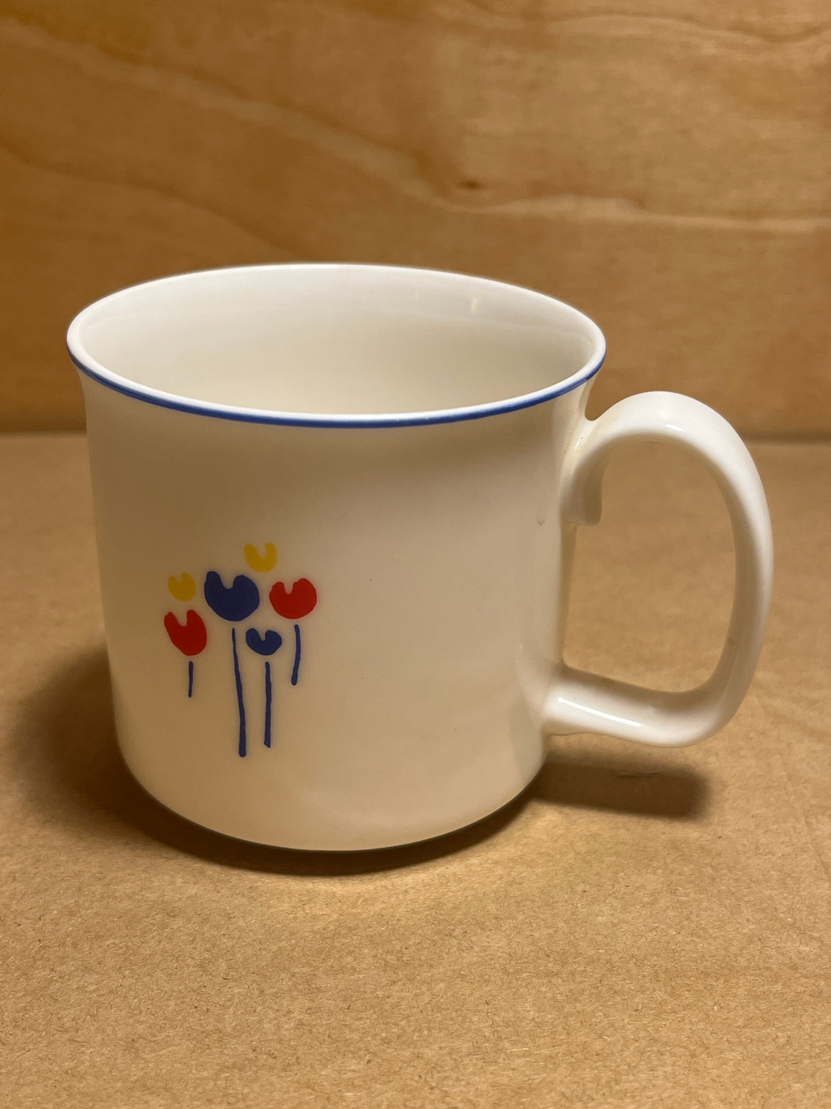 Rival Designs Tulip dinnerware collection Lot 3078 features a white ceramic mug with a blue-edged lip and minimalist floral motif, showcasing tulip-like blooms and blue stems.