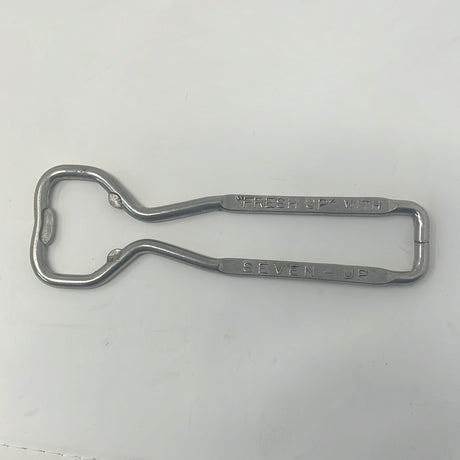 Vintage bottle opener featuring Fresh Up with Seven-Up inscription, alongside tongs and metal sign on white surface.