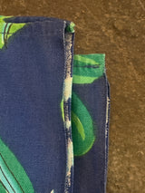 Close-up of a Vintage tablecloth featuring a leafy green pattern on blue fabric, showcasing raw edges and hemmed seams against a stone-like surface.