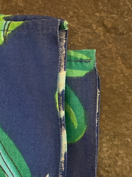 Close-up of a Vintage tablecloth featuring a leafy green pattern on blue fabric, showcasing raw edges and hemmed seams against a stone-like surface.