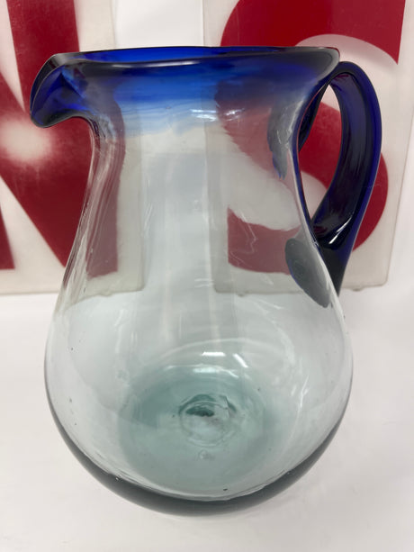 Handblown glass pitcher with blue rim and handle, featuring a wide base for stability and a large handle for easy pouring.