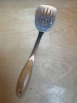 Wolfgang Puck stainless steel perforated spatula with a bamboo handle resting on a light wood surface, ideal for flipping and lifting foods.