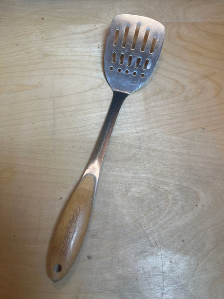 Wolfgang Puck stainless steel perforated spatula with a bamboo handle resting on a light wood surface, ideal for flipping and lifting foods.