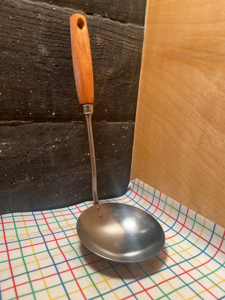 Stainless steel ladle with wood handle - EKCO Eterna, featuring a wide base and durable construction, resting on a patterned cloth.
