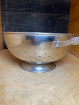 Large colander with handles and elevated base - stainless steel - Sharda, displayed on a wooden shelf, highlighting its sturdy handles and perforated design for effective draining.