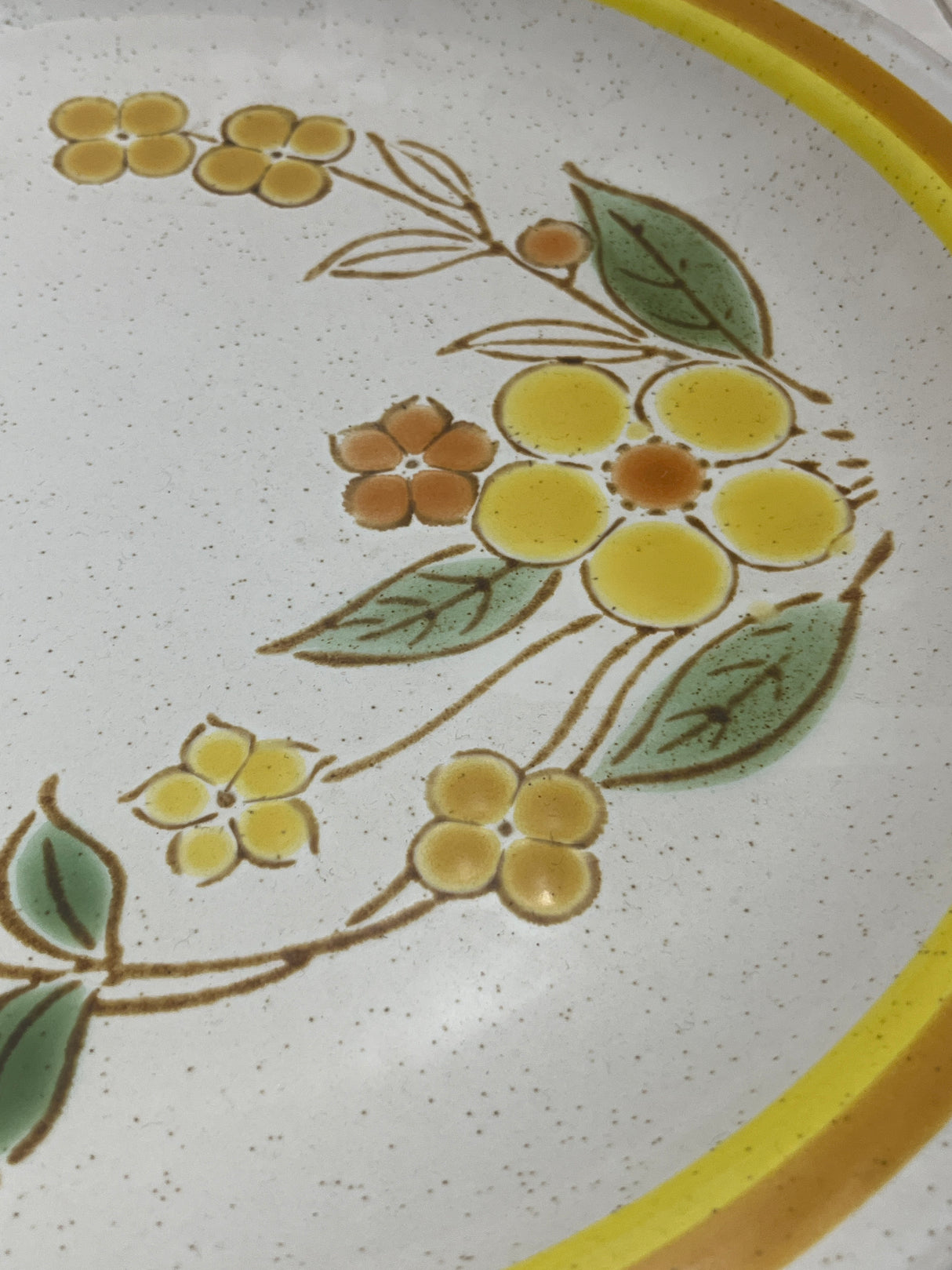 Sunny speckled floral serving plate with a yellow floral motif, showcased with surrounding leaves, highlighting its ceramic ware from International China’s Country Meadow line.