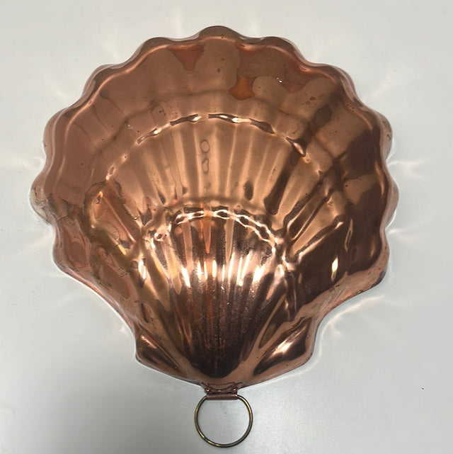 A Large copper seashell baking pan with nickel lining in the shape of a scallop shell with a ring for hanging displayed on a white kitchen surface.