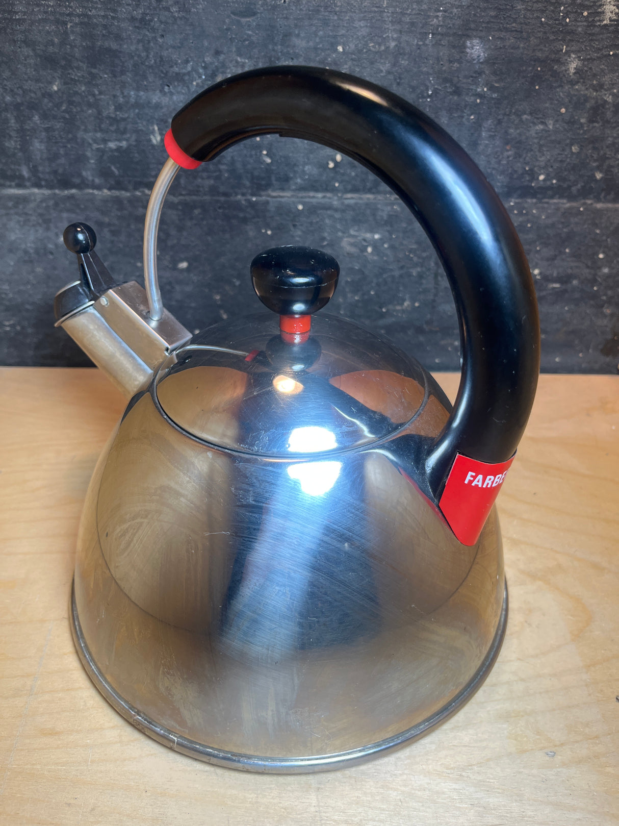 Whistling tea kettle - Farberware 2.5 quart - black handle with red accents, stainless steel body, sits on a wooden surface, featuring a spout lever.