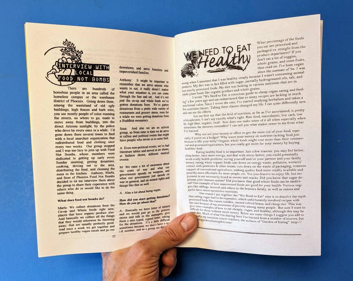Hand holding We Need To Eat! (Zine), featuring interviews on feeding communities and articles on affordable vegan eating, against a blue background.
