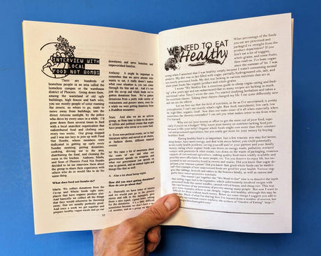 Hand holding We Need To Eat! (Zine), featuring interviews on feeding communities and articles on affordable vegan eating, against a blue background.