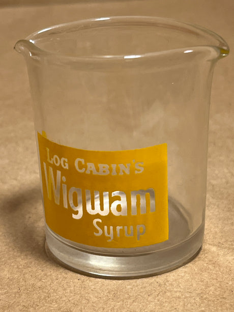 Log Cabin Wigwam syrup pitcher, clear glass with a spout, features a yellow label reading “Log Cabin’s Wigwam Syrup,” designed for easy pouring.