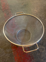 Basket mesh strainer with two handles, made of stainless steel, sits on a stone floor. Features fine mesh for draining pasta and rinsing vegetables.